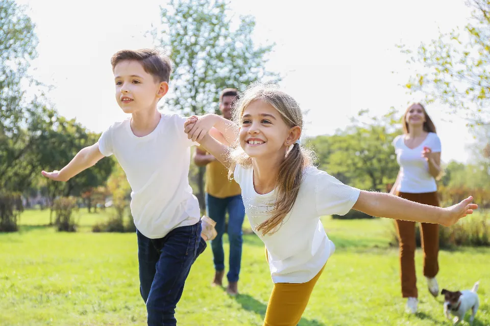 Deux enfants courent dans l'herbe d'un parc en riant, leurs parents derrière eux